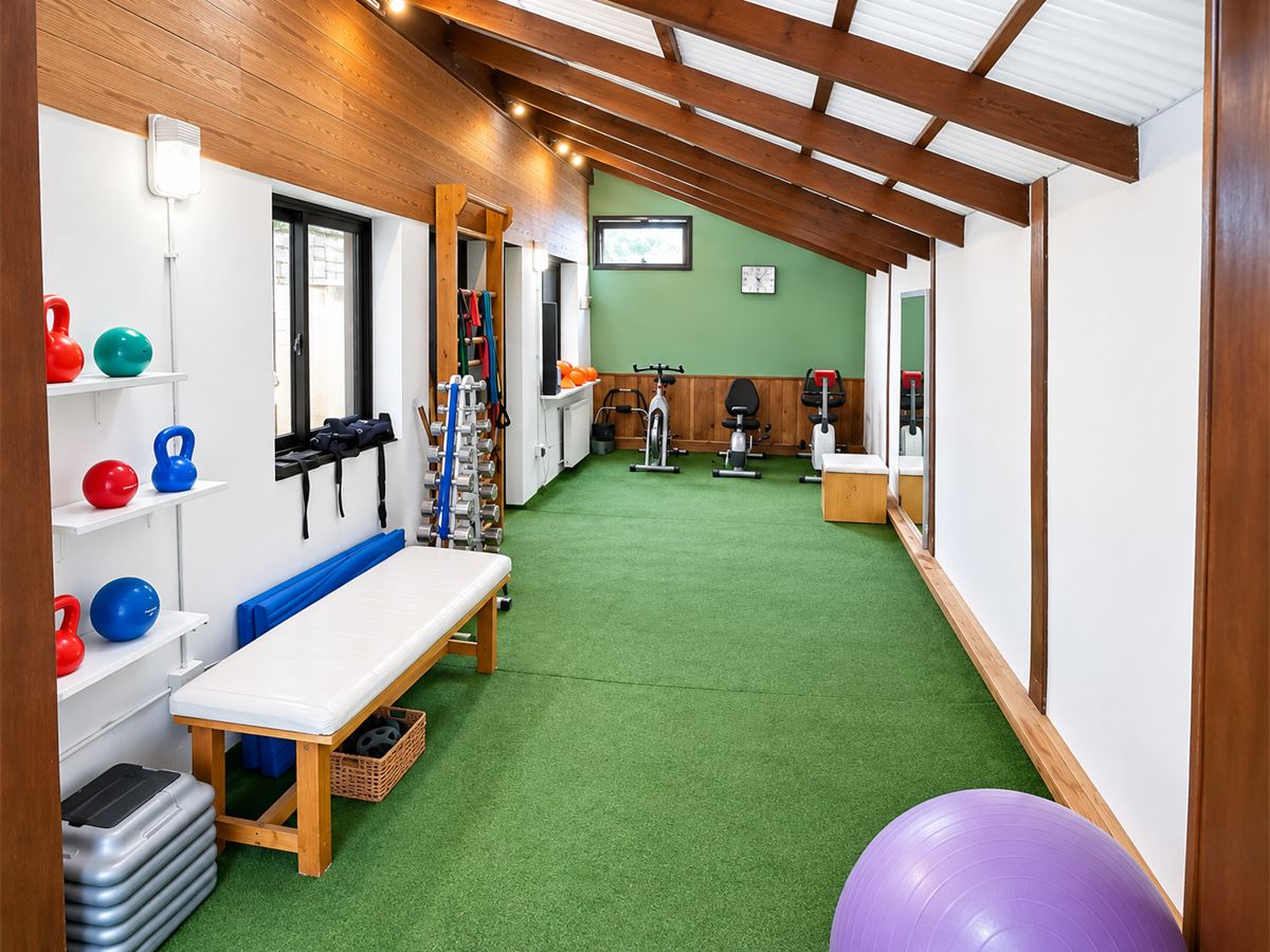 Vista general del gimnasio: piso de césped sintético, banco, pesas rusas, espaldar y bicicletas al fondo